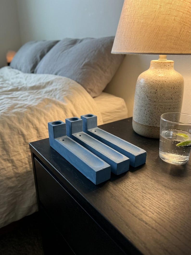 May include: Three rectangular blue concrete incense holders arranged side by side. Each holder features a cylindrical opening at one end and a long, narrow recessed channel extending along the top surface for holding incense sticks.