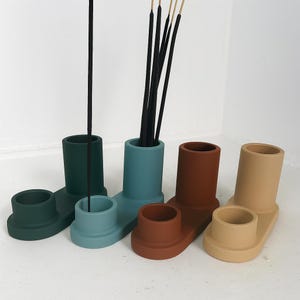 May include: A collection of four ceramic incense holders in various colors: dark green, teal, rust, and beige. Each holder has a tall, cylindrical section for incense sticks and a smaller, round section for catching ash.