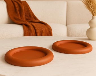 Peut inclure: Deux plateaux ronds de couleur terre cuite avec un bord surélevé reposent sur une surface semblable à du marbre. Une couverture de couleur rouille est drapée sur un canapé blanc en arrière-plan. Un vase avec des plantes séchées est également visible.