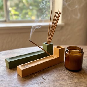 May include: Two rectangular concrete incense holders, one olive green and one mustard yellow, each featuring a cylindrical opening for incense sticks and a long groove. A burning incense stick emits smoke, and a small amber glass candle jar sits nearby.