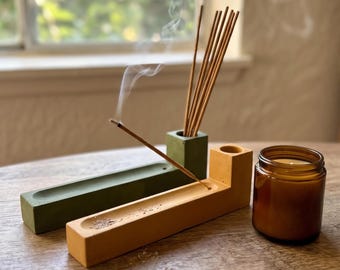 May include: Two rectangular concrete incense holders, one olive green and one mustard yellow, each featuring a cylindrical opening for incense sticks and a long groove. A burning incense stick emits smoke, and a small amber glass candle jar sits nearby.
