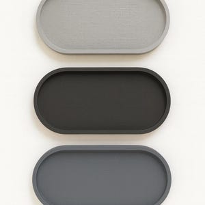 May include: Three oval-shaped trays in a vertical arrangement. The top tray is light gray, the middle one is black, and the bottom tray is dark gray. The trays have a smooth, matte finish and are set against a white background.