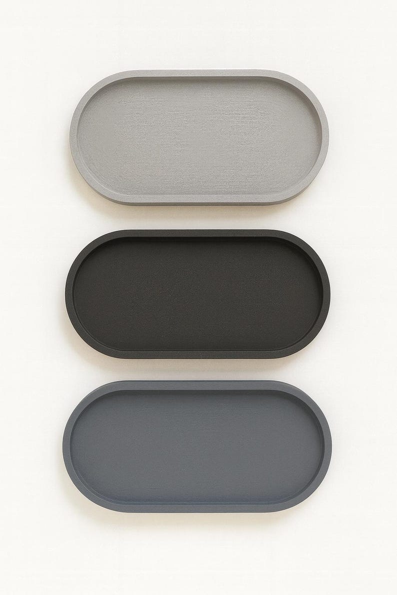 May include: Three oval-shaped trays in a vertical arrangement. The top tray is light gray, the middle one is black, and the bottom tray is dark gray. The trays have a smooth, matte finish and are set against a white background.