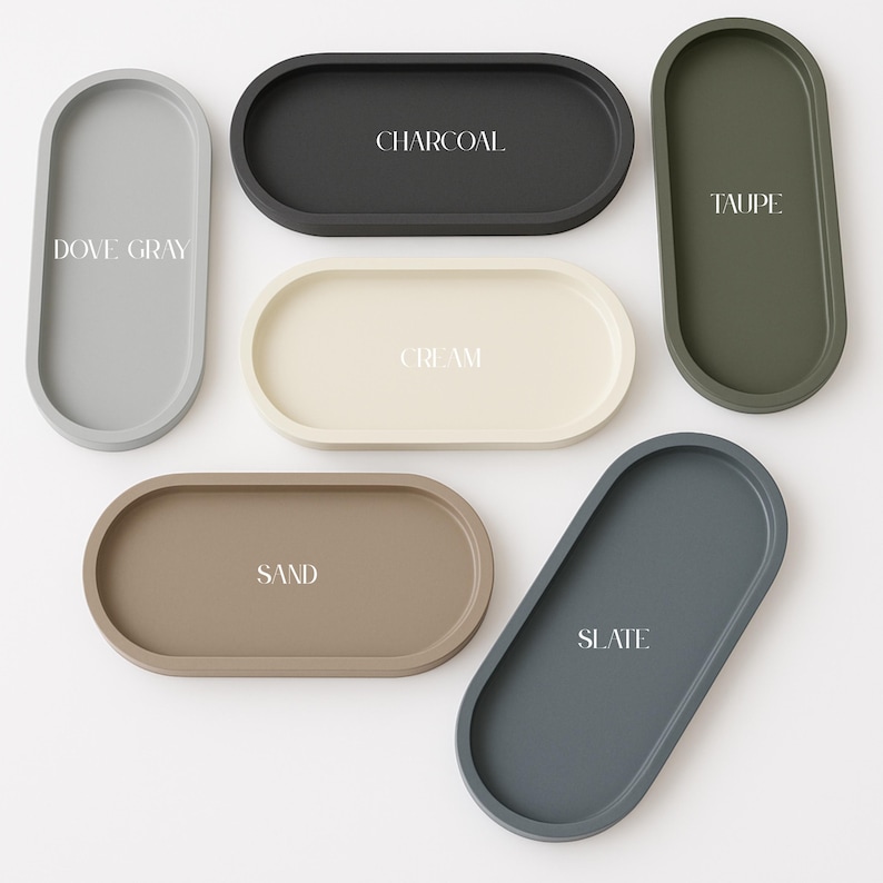May include: An assortment of oval-shaped trays in various colors, including dove gray, charcoal, cream, taupe, sand, and slate. Each tray has a matte finish and features a color name in white text. These trays are ideal for home decor and organization.