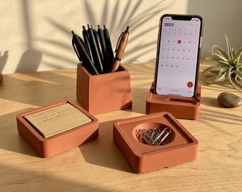 Peut inclure: Un ensemble d'organisateurs de bureau en béton couleur terre cuite comprenant un pot à crayons carré avec des stylos noirs et en bois, un porte-notes carré, un vide-poches rond avec des trombones argentés et un support de téléphone affichant 2021.