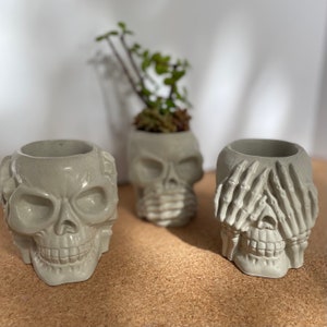 May include: Three gray concrete skull planters. The first skull has its mouth covered, the second has its eyes covered, and the third has its ears covered.
