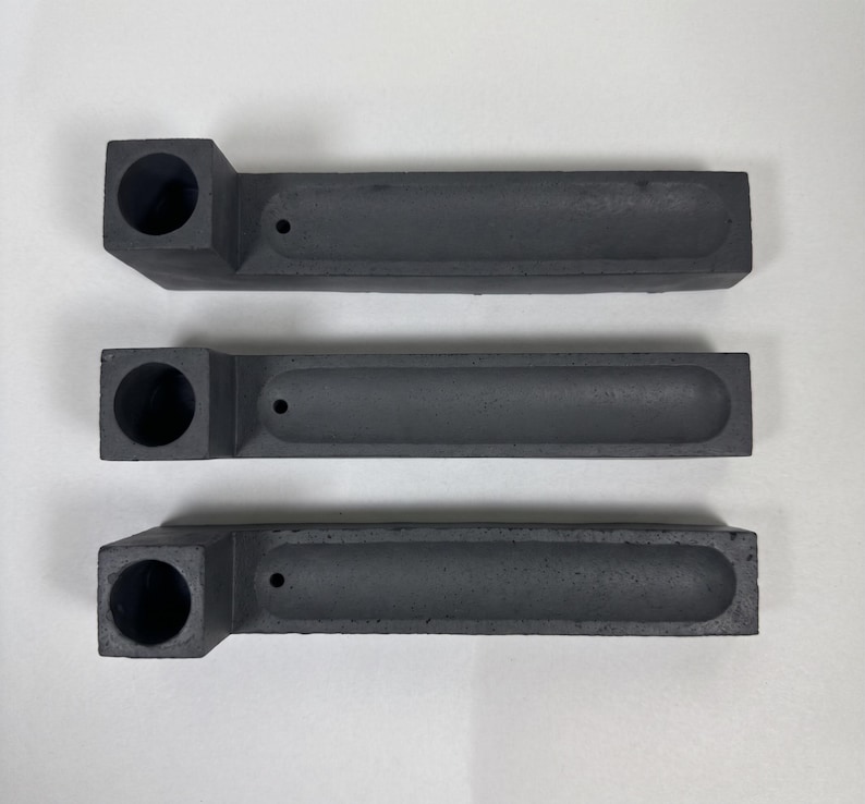 May include: Three dark grey rectangular incense holders arranged horizontally. Each piece features a square block on the left side with a circular opening and a long, narrow, recessed channel extending to the right, ending with a small hole.
