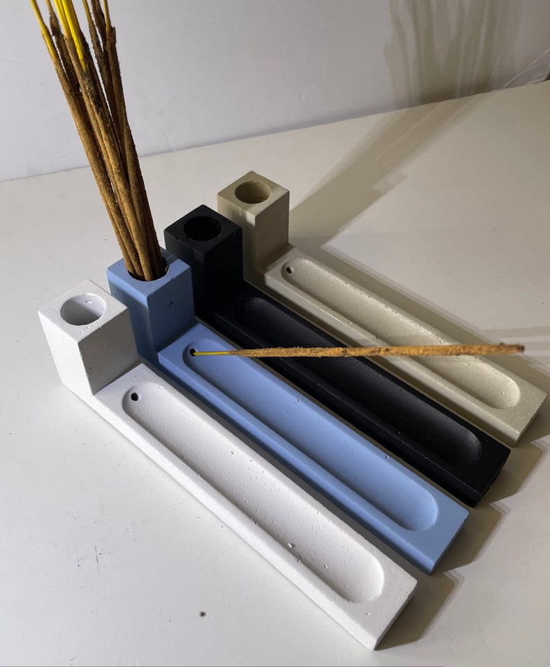 May include: Four rectangular incense holders made of concrete in white, light blue, black, and beige. Each holder features a square block with a cylindrical hole for incense sticks and a long, narrow, recessed tray for catching ash.