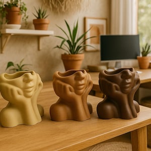 May include: Three ceramic face planters in shades of yellow, brown, and dark brown. The planters have a unique design, with the face resting on the hands. The planters are on a wooden surface.