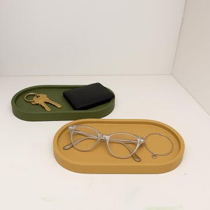 May include: Two oval-shaped trays in olive green and mustard yellow. The green tray holds a set of keys and a black wallet. The yellow tray contains a pair of clear-framed glasses and a gold bracelet. Minimalist design.
