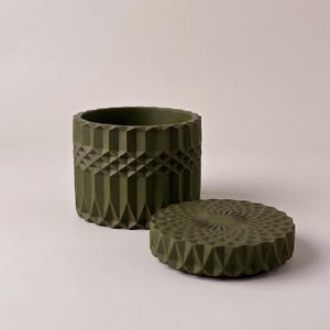 May include: A cylindrical olive green container with a geometric, faceted exterior pattern and a matching circular lid. The lid features a radial, star-like textured design, while the container body displays a repeating vertical diamond motif.