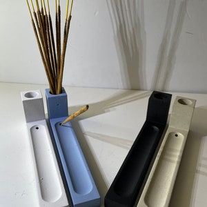 May include: Four L-shaped incense holders in white, light blue, black, and beige. Each holder features a rectangular base with a long, shallow groove and a vertical block at one end containing a circular opening for incense sticks.