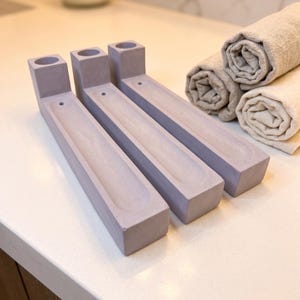 May include: Three light purple, rectangular incense holders made of a smooth, matte material. Each holder features a cylindrical opening at one end for an incense stick and a long, narrow, recessed channel running along the centre of the base.