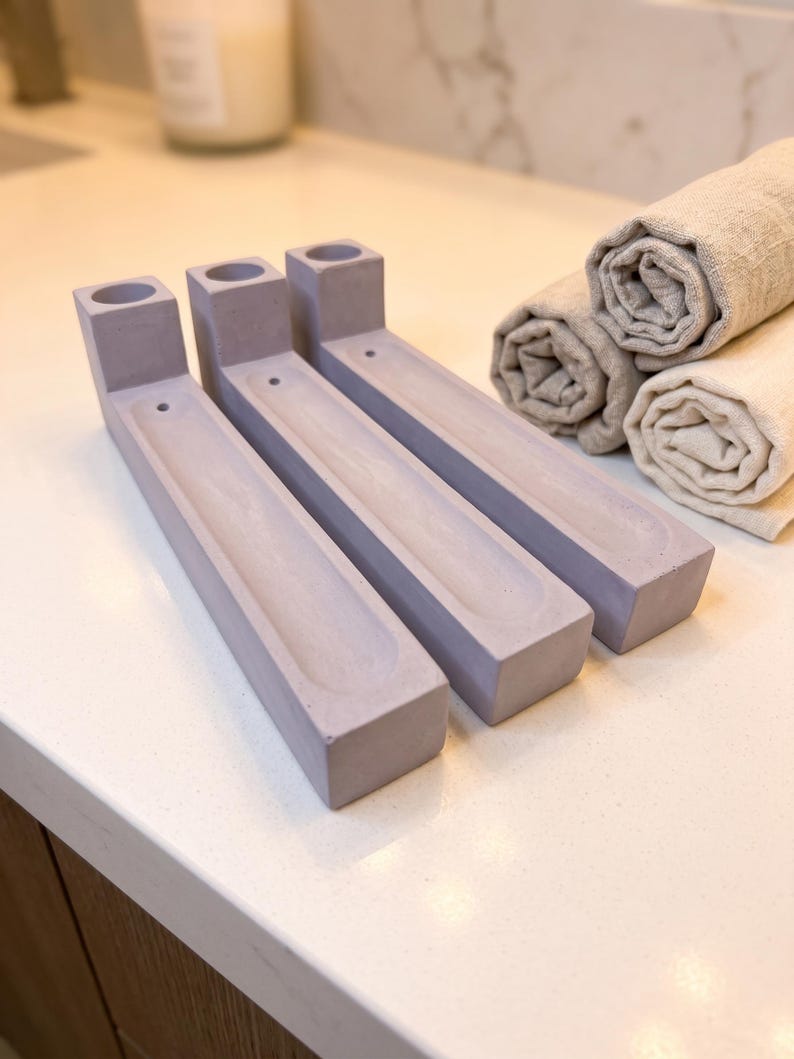 May include: Three light purple, rectangular incense holders made of a smooth, matte material. Each holder features a cylindrical opening at one end for an incense stick and a long, narrow, recessed channel running along the centre of the base.
