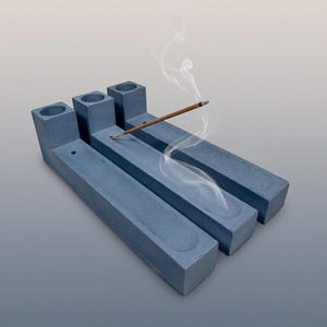 May include: Three rectangular blue-grey concrete incense holders arranged side by side. Each holder features a cylindrical opening at one end and a long, shallow channel running the length of the base. A single lit incense stick rests in one.