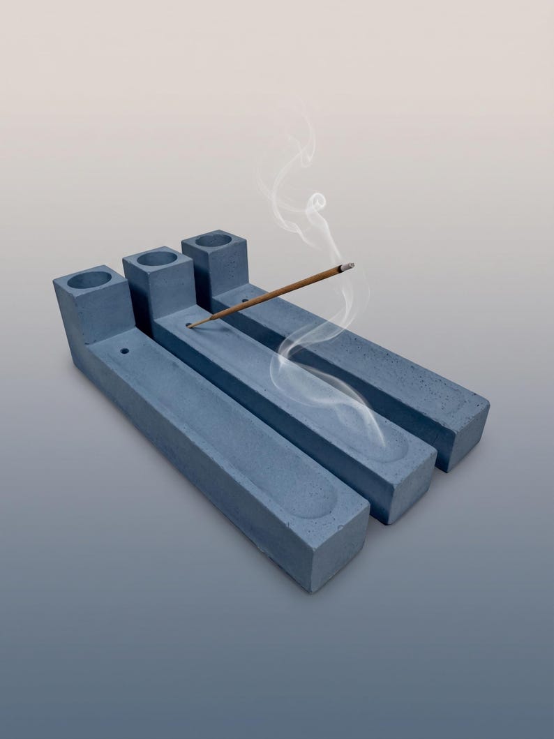 May include: Three rectangular blue-grey concrete incense holders arranged side by side. Each holder features a cylindrical opening at one end and a long, shallow channel running the length of the base. A single lit incense stick rests in one.