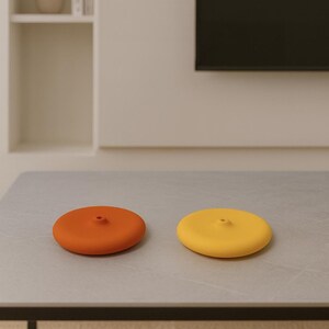 May include: Two circular bottle stoppers, one orange and one yellow. Each has a small central hole. They are displayed on a grey surface. The stoppers are likely made of silicone.
