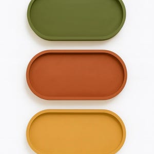 May include: Three oval-shaped trays in olive green, rust orange, and mustard yellow. The trays are arranged vertically against a white background. The trays have a smooth, matte finish and a slightly raised edge, suitable for serving or display.