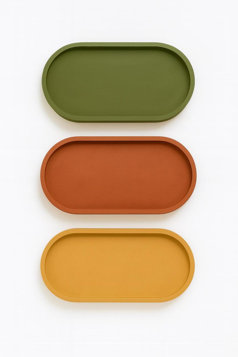 May include: Three oval-shaped trays in olive green, rust orange, and mustard yellow. The trays are arranged vertically against a white background. The trays have a smooth, matte finish and a slightly raised edge, suitable for serving or display.