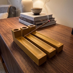 May include: A mustard yellow concrete incense holder featuring three parallel rectangular channels with circular holes at one end. A single lit incense stick is inserted into one of the holes, with a thin trail of smoke rising upward from the glowing tip.