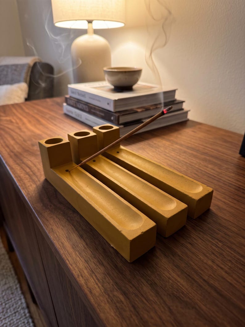 May include: A mustard yellow concrete incense holder featuring three parallel rectangular channels with circular holes at one end. A single lit incense stick is inserted into one of the holes, with a thin trail of smoke rising upward from the glowing tip.