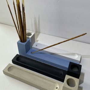 May include: A set of three rectangular concrete incense holders in light blue, black, and beige. Each holder features a cylindrical opening for incense sticks and a long, narrow trough for ash collection. One blue holder contains several incense sticks.