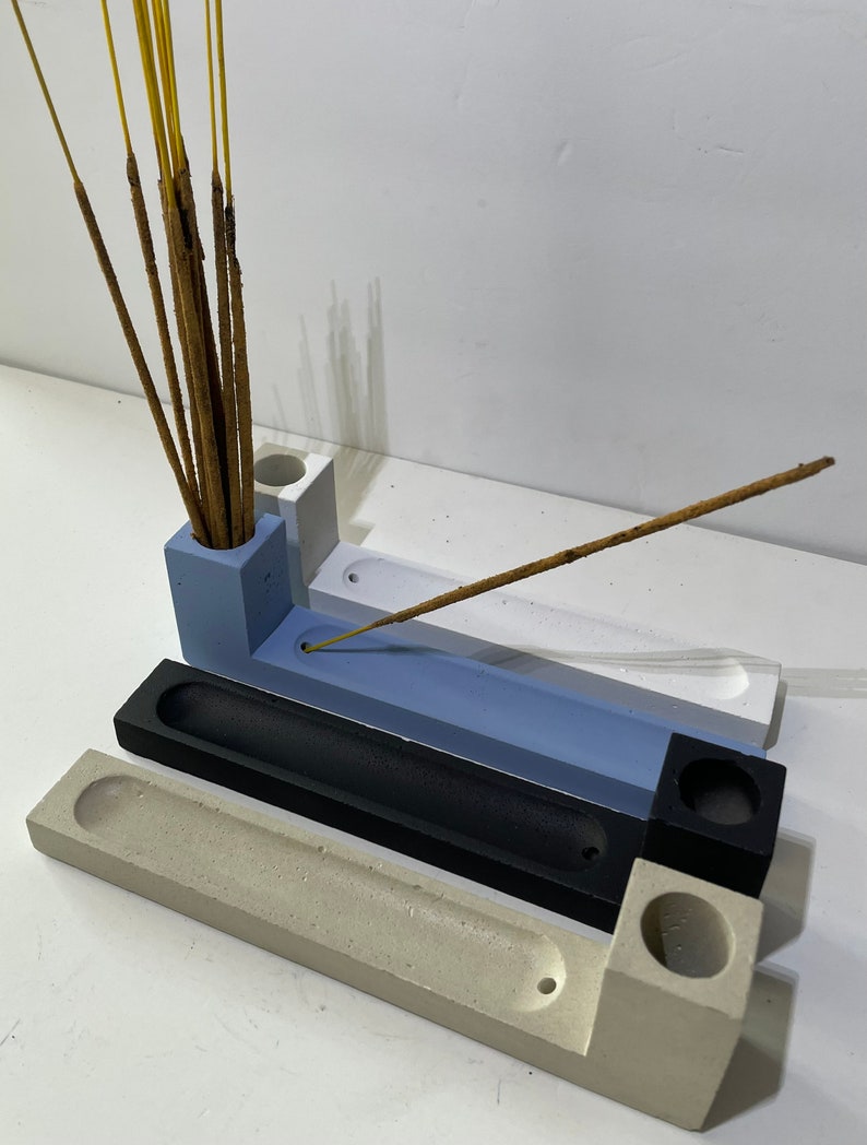 May include: A set of three rectangular concrete incense holders in light blue, black, and beige. Each holder features a cylindrical opening for incense sticks and a long, narrow trough for ash collection. One blue holder contains several incense sticks.