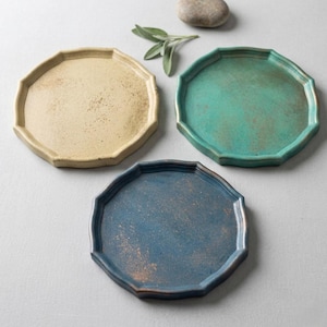 May include: Three decorative ceramic plates in beige, teal, and blue. Each plate has a unique octagonal shape with a textured surface. The plates are arranged on a light gray surface, with a small stone and sprig of greenery.