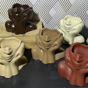 May include: Five ceramic planters shaped like human heads with their hands covering their faces. The planters are in various colors, including brown, beige, and white.