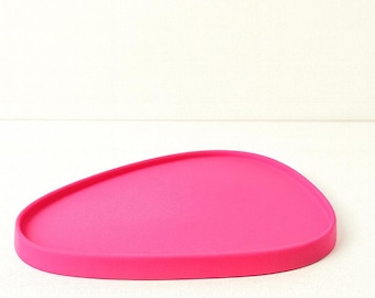 May include: A bright pink, irregularly shaped tray with a slightly raised edge. The tray is made of a solid material and sits on a white surface. The design is simple and modern.