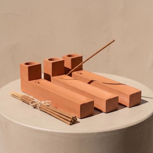 May include: Three terracotta-coloured rectangular incense holders with a square base and a cylindrical opening at one end. Each holder features a long, narrow channel for ash. A single incense stick is inserted into one holder, and a bundle of incense sticks tied with twine rests in front.