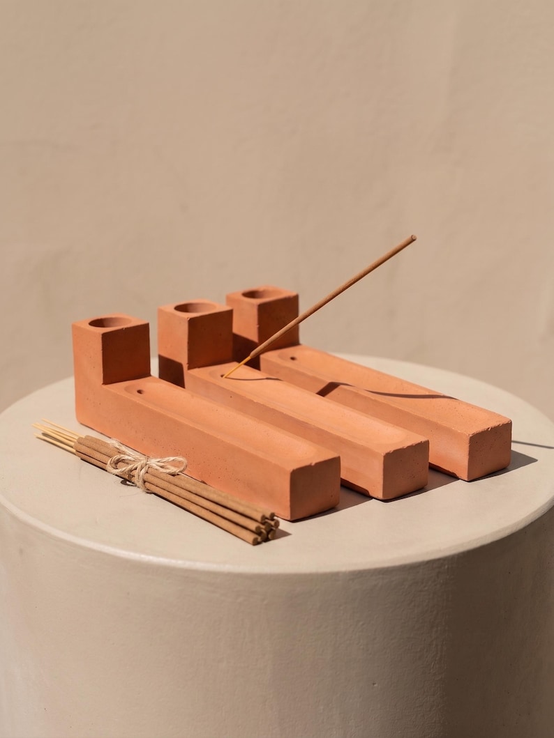 May include: Three terracotta-coloured rectangular incense holders with a square base and a cylindrical opening at one end. Each holder features a long, narrow channel for ash. A single incense stick is inserted into one holder, and a bundle of incense sticks tied with twine rests in front.