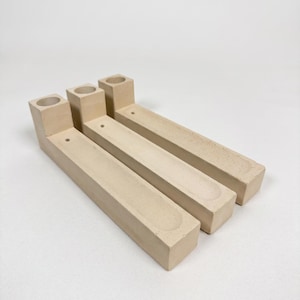 May include: Three beige, L-shaped objects made of a matte, textured material. Each piece features a cylindrical opening at the raised end and a long, shallow, rectangular recessed channel running along the length of the base, with a small hole.