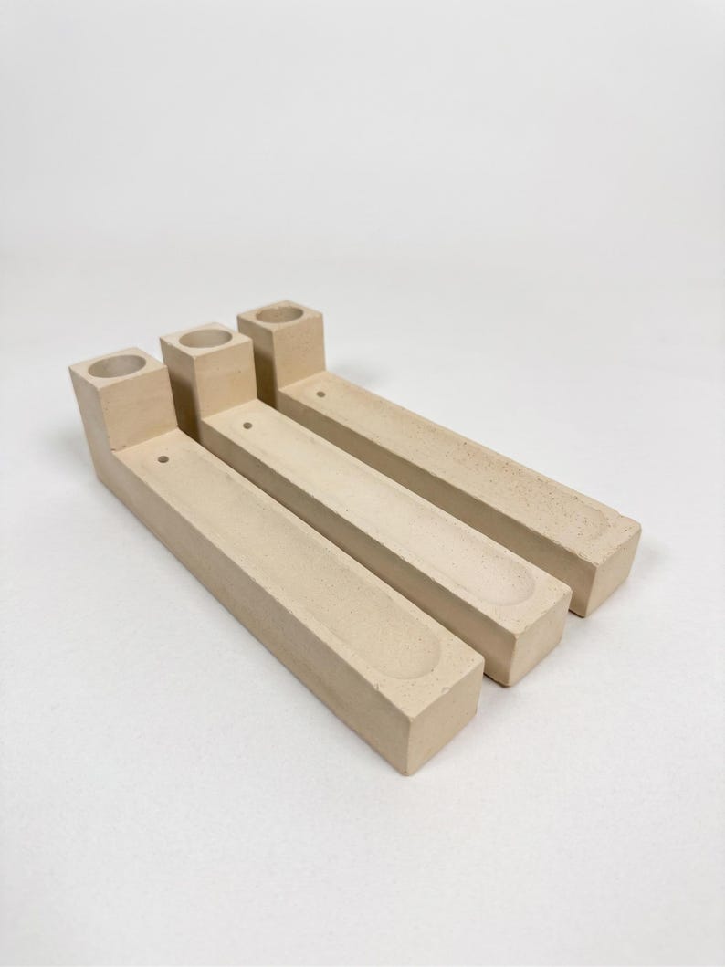 May include: Three beige, L-shaped objects made of a matte, textured material. Each piece features a cylindrical opening at the raised end and a long, shallow, rectangular recessed channel running along the length of the base, with a small hole.