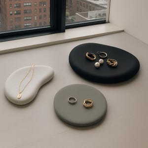 May include: Three jewelry trays in white, black, and gray, each holding various pieces of jewelry. A gold necklace with a pendant rests on the white tray. The black tray displays gold earrings, a ring, and two pearls. The gray tray holds two rings.
