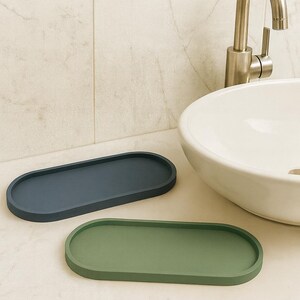May include: Two oval-shaped trays in a bathroom setting. One tray is a muted blue, the other is a muted green. The trays are on a countertop next to a white sink and a silver faucet.