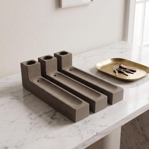 May include: Three rectangular grey concrete candle holders, each featuring a cylindrical opening at one end and a long, narrow recessed tray extending along the base. The holders are arranged side-by-side in a parallel formation on a surface.