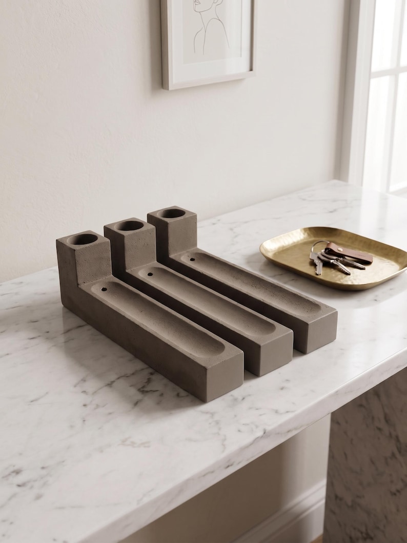 May include: Three rectangular grey concrete candle holders, each featuring a cylindrical opening at one end and a long, narrow recessed tray extending along the base. The holders are arranged side-by-side in a parallel formation on a surface.