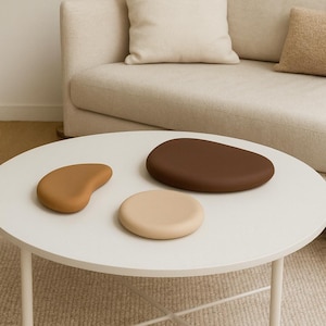 May include: Three decorative stone-shaped objects in shades of brown and beige rest on a white round table. The objects vary in size and shape, with a large, dark brown oval, a smaller beige circle, and a tan, kidney-shaped piece.