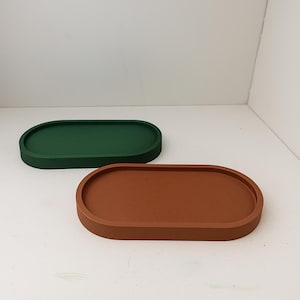 May include: Two oval-shaped trays. One is a deep forest green, and the other is a warm, earthy brown. The trays have a simple, minimalist design with a slightly raised edge. They are likely made of a solid material, perfect for organizing small items.