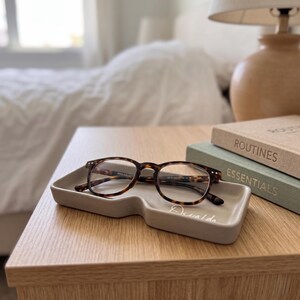 May include: A pair of tortoise shell patterned eyeglasses resting inside a rectangular, light gray ceramic tray. The tray features a small cutout on one side and the word "Oswald" written in white cursive script on the bottom surface.