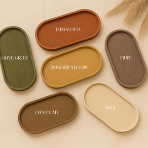 May include: An assortment of oval-shaped trays in various colors, including olive green, terracotta, mustard yellow, taupe, chocolate, and beige. Each tray has a slightly raised edge and features a color name in white text.