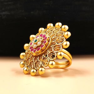 May include: Ornate gold-colored ring with a floral design. The ring features a central cluster of red and green gemstones surrounded by detailed filigree and gold spheres. The ring has a double band.