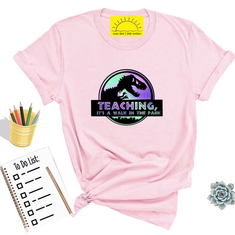 May include: A light pink t-shirt featuring a graphic of a dinosaur silhouette inside a circular design with the text "TEACHING, IT'S A WALK IN THE PARK". The shirt has rolled-up sleeves and a crew neck.