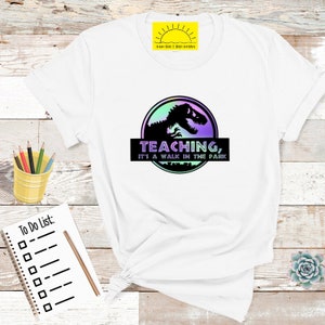May include: White t-shirt with a graphic featuring a dinosaur silhouette inside a circle with a purple and green gradient. The text "TEACHING, IT'S A WALK IN THE PARK" is below the dinosaur image. A yellow pencil holder and a notepad are in the background.