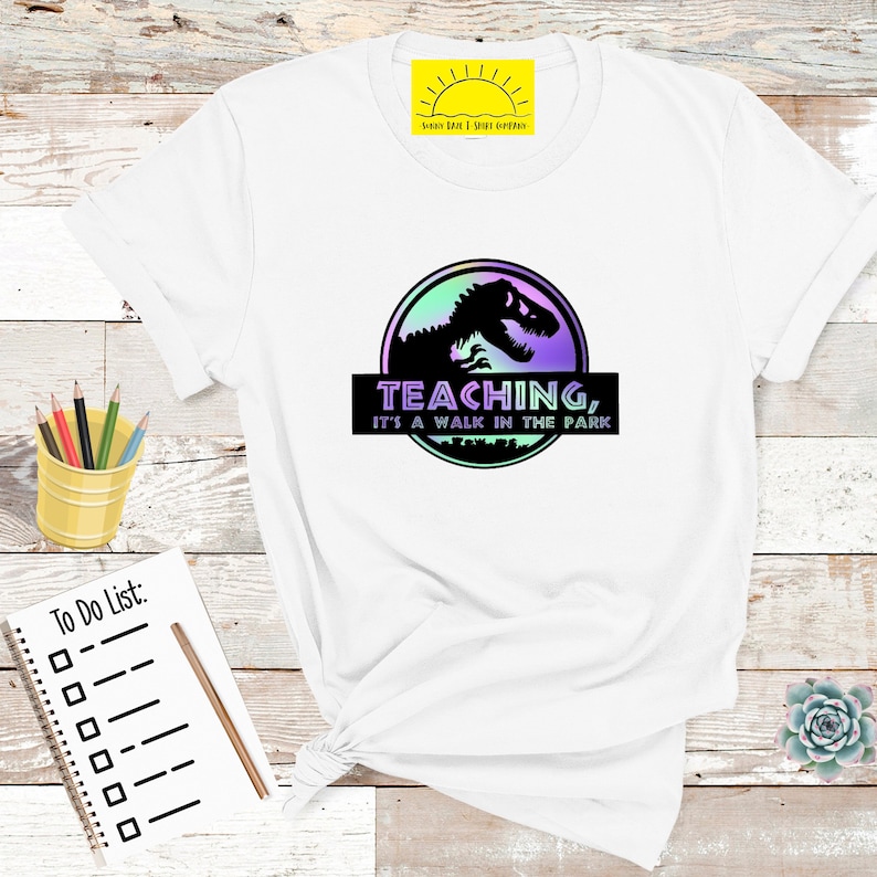 May include: White t-shirt with a graphic featuring a dinosaur silhouette inside a circle with a purple and green gradient. The text "TEACHING, IT'S A WALK IN THE PARK" is below the dinosaur image. A yellow pencil holder and a notepad are in the background.