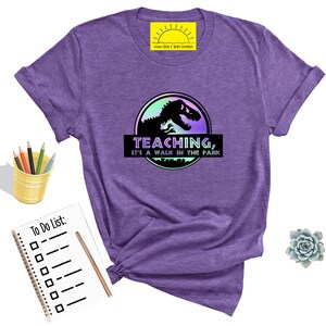 May include: Purple t-shirt with a graphic featuring a dinosaur silhouette inside a circle with the text "TEACHING, IT'S A WALK IN THE PARK." The shirt has rolled-up sleeves and is next to a notepad, pencils, and a succulent.