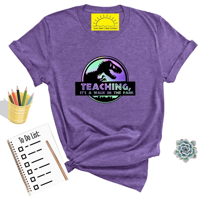 May include: Purple t-shirt with a graphic featuring a dinosaur silhouette inside a circle with the text "TEACHING, IT'S A WALK IN THE PARK." The shirt has rolled-up sleeves and is next to a notepad, pencils, and a succulent.