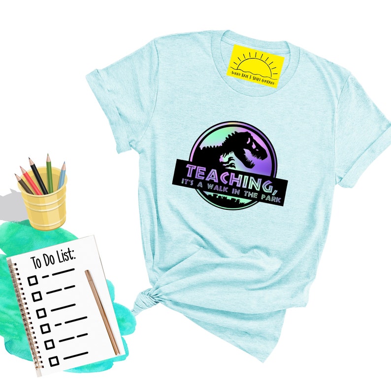 May include: Light blue t-shirt with a graphic featuring a dinosaur silhouette within a circular design, with the text "TEACHING, it's a walk in the park." A notepad with a to-do list and pencils in a yellow bucket are also visible.