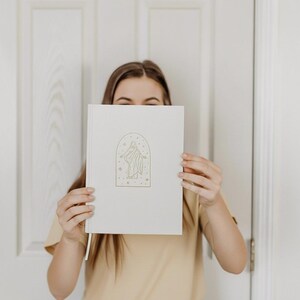 Pu&ograve; includere: Un diario color crema con un'illustrazione dorata di una figura all'interno di una cornice ad arco, circondata da stelle e punti. Il diario ha un segnalibro a nastro abbinato. La copertina &egrave; liscia e il design &egrave; minimalista.